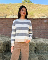 MAGLIONE A RIGHE ANTRACITE E PANNA indossato in un ambiente all'aperto con fieno e natura sullo sfondo.