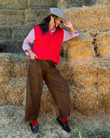 Model posing in hay with red vest, brown PANTALONE BAGGY IN ECOLEATHER BROWN, and stylish cap.