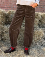 PANTALONE BAGGY IN VELLUTO CASTAGNO indossato su sfondo di paglia con maglione e scarpe eleganti.
