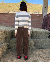 PANTALONE BAGGY IN VELLUTO CASTAGNO worn by a person in striped sweater, standing in front of hay bales.