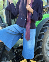 Modella che indossa giacca crop doppiopetto sofisticata con cravatta, pantaloni in denim, accanto a un trattore.